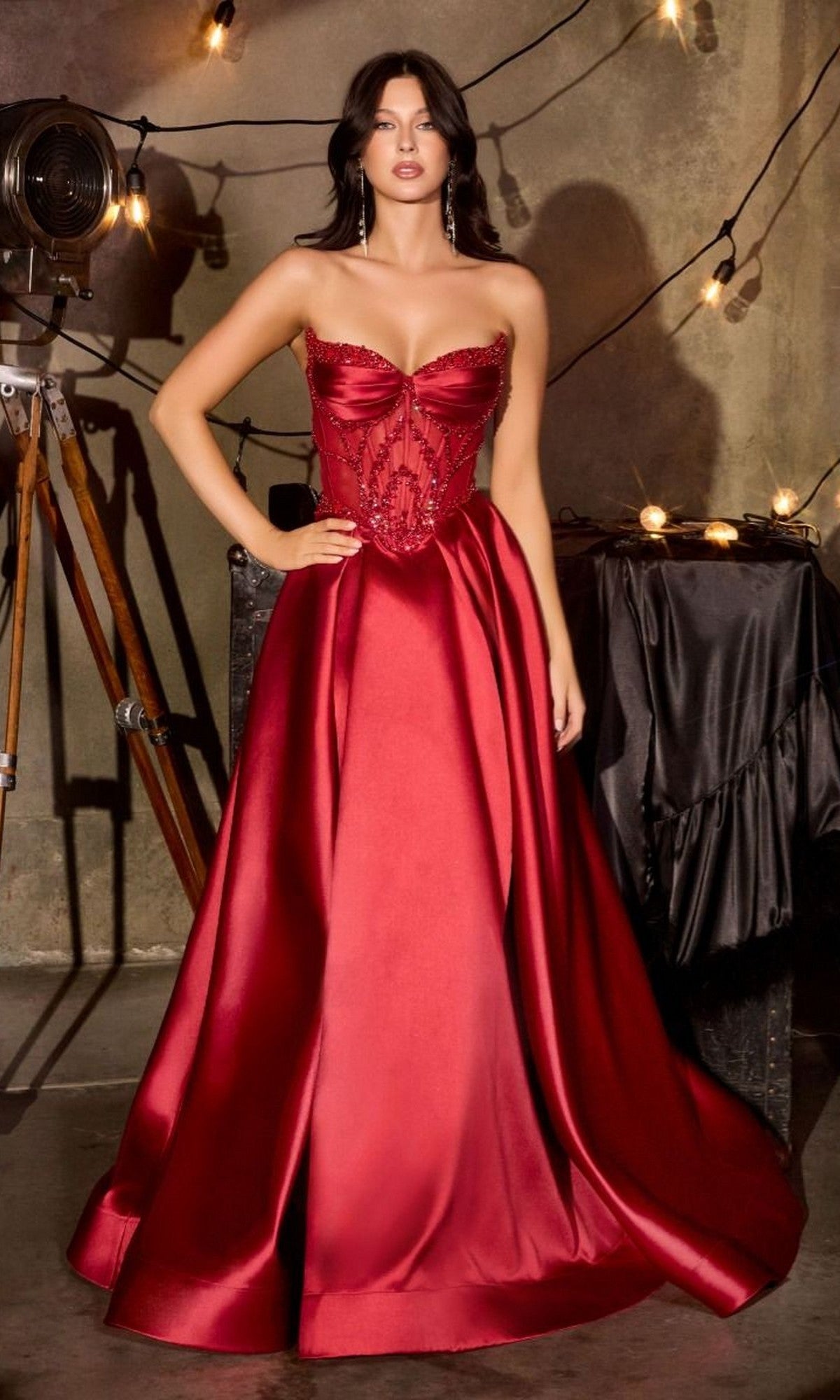 A woman wearing the burgundy Ladivine CD389 long prom dress, featuring a strapless mikado bodice and A-line skirt, stands indoors near string lights and a vintage film camera.